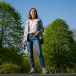 Mladá žena venku na jaře v bundě, bílé blůze a džínách drží sluneční brýle u zelených stromů.